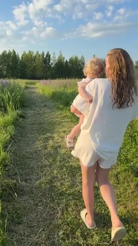 A young woman with long, wavy brown hair is carrying a baby with blonde hair while walking down a dirt path through a grassy field. The woman is wearing a white t-shirt and white shorts. The baby is wearing a white dress. The field is full of green grass and some purple flowers. In the background, there is a line of trees under a blue sky with white clouds. The sun is shining, creating a warm and inviting atmosphere. The woman and baby appear to be enjoying a leisurely stroll through nature.