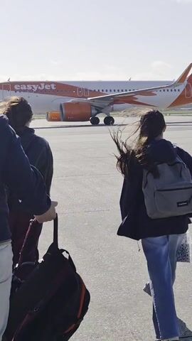 The video shows a group of people walking on an airport tarmac toward an EasyJet passenger plane. A tow vehicle is attached to the plane's front wheel. The plane is white with orange lettering that says "easyJet" on the fuselage. There are 3 people in the foreground, with the person on the left wearing off-white pants and a blue shirt. The person in the center is wearing burgundy pants and a dark shirt, and the person on the right is wearing jeans and a black sweatshirt with a gray backpack.  The sky is blue and the ground is gray asphalt.