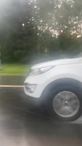 This video shows a view from inside a vehicle driving on a wet road during a rain shower. The road surface is dark and reflective, with white lines indicating lanes. On the right side of the road, there is a shoulder of green grass followed by a line of trees. In the distance, a few mile markers can be seen. The side mirror of the vehicle is visible on the left side of the frame, covered in raindrops. The sky appears overcast and bright. The camera is moving, indicating the vehicle is in motion.
