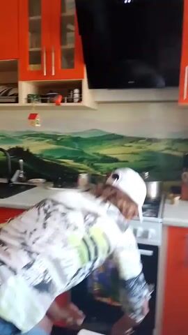 A woman wearing a white baseball cap, a patterned shirt, and denim shorts is in a kitchen. She is bending over a stovetop with a pot on it, and then moves to a counter where she picks up a pan. She then turns to face the camera, with a slight smile, and says "I'll tell you when." The kitchen has orange cabinets and a backsplash with a landscape mural.
