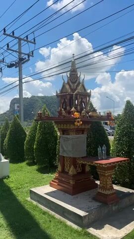A small, ornate structure, possibly a shrine or spirit house, stands on a concrete base in a grassy area. The structure is elaborately decorated with red and gold accents and features a multi-tiered, pagoda-like roof. A grey cloth is draped around the central pillar. To the right of the shrine, a small red table with gold trim holds two bottles of water. The shrine is flanked by rows of neatly trimmed green conical trees. In the background, a mountain is visible under a bright blue sky with scattered white clouds. Power lines stretch across the top of the frame. The scene appears to be outdoors, possibly by a roadside, given the presence of power lines and the style of landscaping. The lighting suggests it is daytime, with clear weather. There are no people or animals visible in the frame.