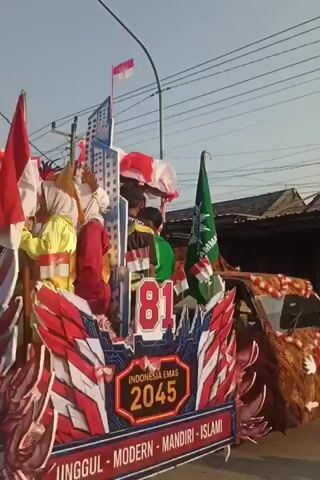 The video shows a parade float decorated with red, white, and blue motifs, featuring an Indonesian flag at the top. Several people are on the float, including a child dressed in traditional attire and a person in a green shirt. A banner on the float reads "INDONESIA EMAS 2045" and "UNGGUL - MODERN - MANDIRI - ISLAMI". To the right of the float is a truck decorated with a brown batik-like pattern, with people inside. The truck also has elaborate, colorful decorations resembling a stylized bird or dragon head on its side. Another float is visible behind the truck, decorated with similar colorful patterns and flags. The parade appears to be taking place on a street with power lines and buildings in the background, under a clear sky, possibly during the day.