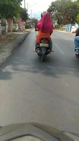 The video shows a first-person perspective of someone riding a motorcycle on a paved road. The rider is following two other motorcycles. The first motorcycle ahead has two people on it, a driver and a passenger wearing an orange shirt and a red hijab. The second motorcycle ahead is ridden by a single person wearing a backpack. The road is lined with buildings on the left and right sides, with some trees and utility poles visible. The sky is overcast. The video appears to be taken during daylight hours.