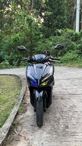 The video shows a black motorcycle parked on a paved road. The motorcycle has illuminated headlights, with the left side emitting a blue light and the right side emitting a yellow light. The motorcycle is positioned centrally in the frame, facing the viewer. The background consists of lush green foliage and trees, with a slightly overcast sky. The road appears to be curving to the left, and a concrete curb is visible on the left side of the road. The lighting suggests it is either early morning or late afternoon. There are no people visible in the video.