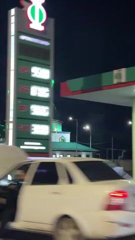 The video shows a mosque illuminated by green lights at night. The mosque has two tall minarets and a large green dome. There are smaller domes on the roof as well. The building is white with green accents. The lights outline the architectural features of the mosque. In the foreground, there is some blurry greenery and a roadside. To the right, a tall structure with illuminated panels is partially visible. The video appears to be filmed from a moving vehicle.