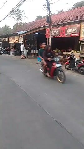A video shows a street with several shops and motorcycles. In the foreground, a person is riding a motorcycle and then dismounts to enter a shop. Another person is seen standing outside a shop, and several motorcycles are parked in front of the shops. The shops appear to be selling goods such as food and drinks. The street is paved, and there are trees and buildings along the sides. The sky is clear, and the lighting suggests it is daytime.