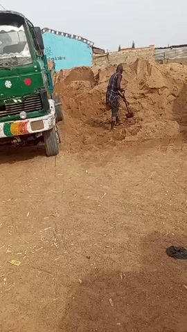 A man is shoveling sand next to a green truck. The man is wearing a plaid shirt and dark pants. He is standing on a large pile of sand and using a shovel to scoop up the sand. The truck is parked next to the pile of sand. The ground is dirt. There is a blue building in the background.