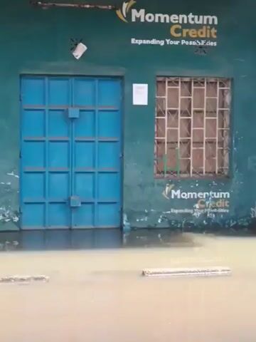 The video shows a street scene, mostly flooded with standing water. The water level covers about half the height of the doors to a row of businesses, including a Momentum Credit and DStv shop. The building is one story, with barred windows. The lighting is bright, suggesting it is daytime, and the video appears to be taken from street level. The color of the water suggests it may be dirty or silty. There is a wooden table near the DStv shop door.