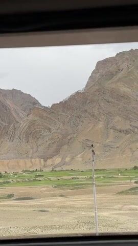 The video shows a view from inside a vehicle looking out at a mountainous landscape. The mountains are rugged and arid with visible layers of rock formations, some of which appear folded. The sky is overcast. In the middle ground, there is a strip of green farmland or vegetation, with a few small structures that could be buildings or tents. The foreground is blurry and appears to be part of the vehicle's interior, possibly a window frame or dashboard. A dark, circular object, possibly a camera lens or part of the vehicle, is visible in the lower left corner of the frame at the beginning of the video. As the video progresses, the view pans slightly to the right, revealing more of the mountainous terrain and the green valley.