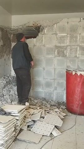 A man is using a power drill to break apart a wall made of glass blocks. The man is wearing a dark jacket, pants, and hat, and is standing amid piles of debris, including broken tiles and chunks of concrete.  The glass block wall is mostly intact except for a hole near the top, and the room appears to be undergoing renovation or demolition. A large red cylindrical object stands next to the wall. The surrounding walls are also damaged, and there is exposed rebar visible in the ceiling.
