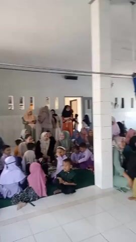 The video shows a group of people, primarily women and children, gathered inside a building that appears to be a mosque or religious center. The room is decorated with various displays resembling tiered structures, adorned with packaged food items such as snacks and drinks, and some fabric decorations. The walls are painted with a sky-like mural, and there are ceiling fans visible. Some individuals are seated on the floor, while others are standing. The atmosphere seems to be a gathering or event.