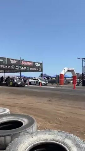 The video shows a drag race with two cars starting from the same line, one a dark grey Mini Cooper and the other a white sports car. The drag race is on a dry dirt track with tires stacked to the side to create a barrier. In the background, there are tents and people watching the race on a sunny day. At the end of the track is an archway and a control tower. The white sports car wins the race.