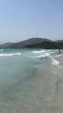 This is a vertical video of a beach on a clear, sunny day. The ocean is a clear turquoise color with gentle waves rolling onto a sandy shore. In the distance, there are green, hilly mountains under a bright blue sky. There are several people visible in the water, some swimming and others wading. One person in a red shirt is standing on the beach in the right side of the frame. The water appears shallow near the shore, and the sandy bottom is visible through the clear water.