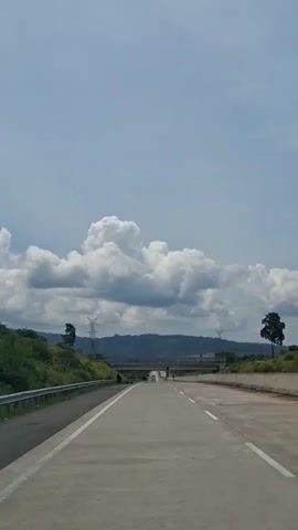 The video shows a highway on a clear day. There are clouds in the sky. There are trees and hills on either side of the road. There are power lines in the distance. There are a few cars on the road. There are also a few people walking on the side of the road. The video is shot from a car.