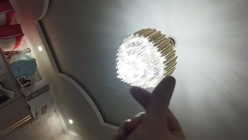 The video shows a white ceiling with a round crystal chandelier with gold accents. A hand is making a heart symbol in front of the chandelier. Part of a room is visible in the lower left corner of the frame, showing a built-in cabinet with items on shelves, and a patterned wall.