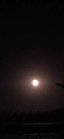 A full moon is visible in the night sky, illuminated by its own light. The moon is centered in the frame, with a soft glow surrounding it. The sky is dark, suggesting it is nighttime. The bottom of the frame shows a dark silhouette of trees or bushes, possibly indicating a landscape. A faint hint of a tree branch or similar object is visible on the right side of the frame, also silhouetted against the sky. The video appears to be a static shot of the moon.
