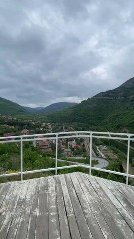 The video shows a scenic viewpoint from a wooden platform with a white metal railing, overlooking a lush valley landscape. The sky is overcast with grey clouds. In the foreground, the wooden platform appears aged with weathered paint. A white railing surrounds the platform's edge, offering a safety barrier while allowing an unobstructed view of the scenery. The valley below features a small town nestled amongst the greenery, with houses having reddish-brown roofs. A road winds through the valley, and a railway track runs alongside it. The hills are covered with dense green forests, and mountain peaks can be seen in the distance, partially obscured by the low-hanging clouds. The overall impression is one of tranquility, despite the cloudy weather.