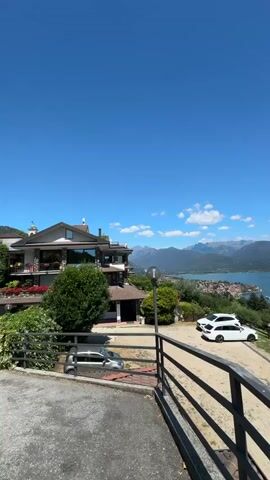 This is a vertical, POV video shot from a high vantage point overlooking a large lake and distant mountains. The camera pans slowly to the right. In the foreground, a grey car is parked on the left, and two white cars, one with a roof rack, are parked on the right. A dark railing runs along the bottom of the frame. Behind the cars, a multi-story building with a brown roof and balconies is visible, surrounded by greenery. Beyond the building, the expansive blue lake stretches out, dotted with small islands and coastal towns. The sky is a clear, bright blue with a few scattered white clouds. The overall scene suggests a scenic vacation spot on a sunny day.