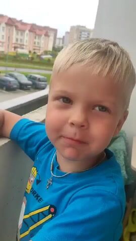 A close-up shot shows a young boy with blond hair wearing a blue t-shirt. He is smiling and looking towards the camera. In the background, there is a balcony with a railing and a view of buildings and cars. The boy is leaning on the railing.