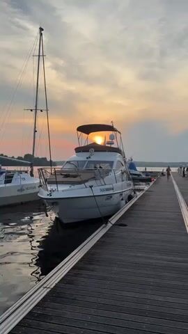 The video opens with a wide shot of a marina at sunset. Several boats are docked, including a large white motor yacht in the foreground. A tall sailboat is visible to the left of the motor yacht. The sun is setting behind the motor yacht, casting a warm orange glow across the sky and reflecting on the water. A wooden pier extends into the water from the right side of the frame. A person in a red dress is walking away from the camera on the pier. The camera then zooms in on the motor yacht, focusing on its canopy and the sun shining through it. The video ends with a close-up of the yacht's equipment and the setting sun.