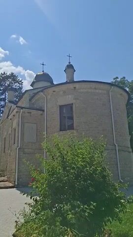 The video begins with a view of a stone church with a dome and a cross on top, surrounded by trees and a blue sky with clouds. In the next clip, a group of men walk past the church. Then, a bell tower is shown with a cross on top. Another clip shows a closer view of the church building with its domes and windows. The video concludes with a view of the church surrounded by trees and grass, with two children in the foreground.