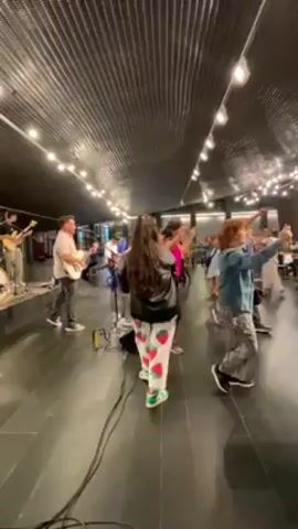 A band performs for a crowd of people. The band consists of a drummer, a bassist, and at least two guitarists. The audience members are facing the band and dancing to the music. The room has a black tiled floor and an undulating dark ceiling with string lights hanging from it.