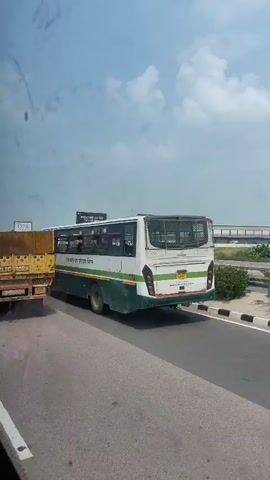 This video shows a traffic scene on a highway. In the foreground, a yellow truck is partially visible on the left. To its right, a white and green bus is traveling on the road. The bus appears to be a public transport vehicle with windows showing passengers inside. The bus has text written on its side in a foreign language, likely Hindi, and a green stripe runs along its bottom. The background shows a highway with a sky filled with clouds. The video is shaky, suggesting it was filmed from a moving vehicle.