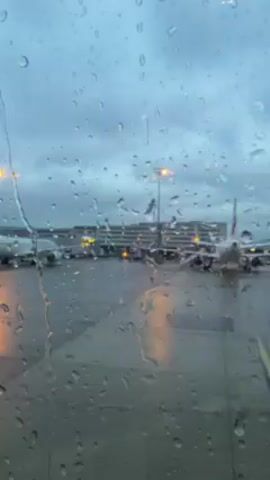 The video shows a view of an airport apron through a window covered in raindrops. The sky is overcast and the light is dim, suggesting a cloudy or rainy day. Several airplanes are visible in the background, along with terminal buildings and lit-up poles. The raindrops on the window obscure the details, creating a blurry effect and accentuating the wet and dreary weather conditions.