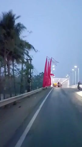 The video shows a view from a moving vehicle on a road. On the left side of the road, there are tall red flags attached to poles. On the right side, there is a bridge with streetlights. A few vehicles are visible on the bridge in the distance, including what appears to be a red truck. The sky is overcast and appears to be either early morning or late evening. The flags are long and flowing, suggesting a windy condition. Some of the flags have text on them, but it is not clearly legible. In the background on the bridge, there is an Indonesian flag.