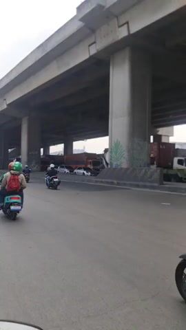 The video shows a view from a vehicle on a road under a concrete overpass. Several motorcycles are visible on the road, including a blue scooter in the foreground with a rider wearing a green helmet, a black scooter behind it with a rider wearing a white helmet, and another black scooter further back with a rider wearing a grey hoodie and a black helmet. In the distance, a large white tanker truck appears to be stopped, possibly due to an accident, along with other trucks and cars. Graffiti is visible on one of the overpass support pillars. The overall scene suggests traffic congestion under an elevated highway.