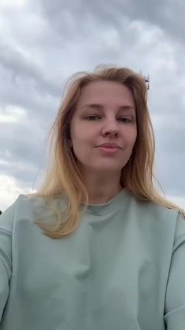 A young woman with blonde hair and freckles, wearing a light blue sweatshirt, is outside with a cloudy sky. In the background, there is a white building with a dark dome and a tall red and white communication tower. The woman smiles and winks at the camera.