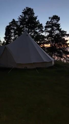 The video shows a sunrise over a body of water, possibly a lake or ocean, as seen from a grassy area with trees and tents. The sky is a gradient of blue, purple, and orange as the sun rises, casting a warm glow over the water. A white bell tent is visible on the left side of the frame, partially obscured by trees. As the camera pans to the right, more trees become visible, silhouetted against the vibrant sky. The water reflects the colorful sky, creating a serene and picturesque scene. No people are visible in the video.