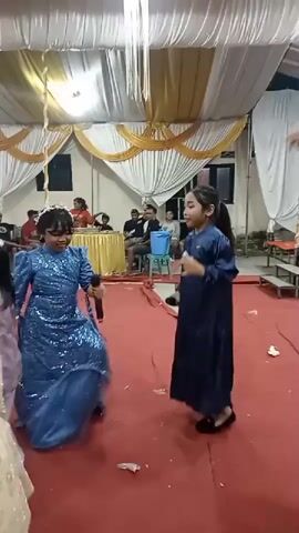 The video shows a group of children and adults at an indoor event, likely a party or reception, indicated by the draped white and gold fabric ceiling and red carpeted floor. In the foreground, three young girls are the main focus. One girl in a sparkly blue dress is holding a microphone and dancing. To her right, another girl in a dark blue dress is also holding a microphone and observing the first girl. To the left of the girl in blue, a third girl in a light-colored dress with a detailed pattern on the bodice is facing away from the camera, also appearing to be dancing. In the background, several adults are seated or standing, some watching the children. The overall atmosphere seems festive.