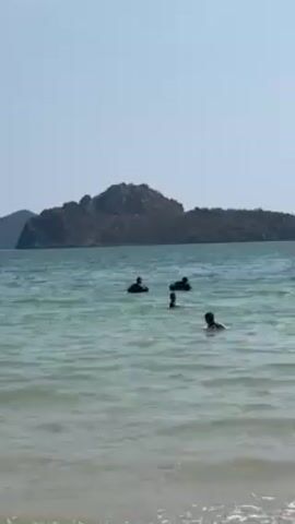 The video shows a calm ocean beach with an island visible in the background. Several people are swimming in the water. A boy wearing a patterned shirt and shorts walks into the water, kicks a blue soccer ball, and plays in the surf.