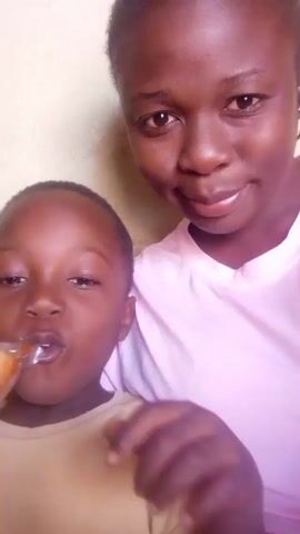 A woman and a boy are posing for a selfie. The woman is behind the boy, with her arm around him. She is wearing a pink shirt. The boy is wearing a beige shirt and holding a small plastic bag filled with a brown substance. The woman holds the bag over the boy's head. The background is a plain, light yellow wall. There is music playing in the background.