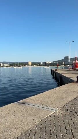 The video shows a view of a harbor with several sailboats anchored in the water. The water is calm and blue, reflecting the clear sky above. In the background, a city skyline with buildings and trees can be seen. The foreground features a concrete pier with textured paving stones. A lamppost is visible on the right side of the pier. The overall scene is serene and depicts a coastal urban environment.