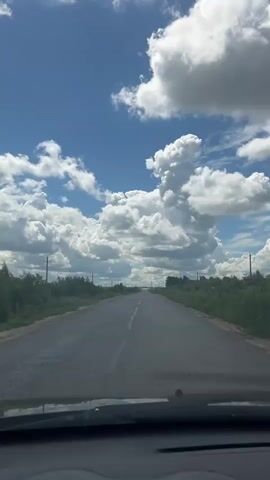 The video shows a POV shot from a vehicle traveling on a paved road. The sky is bright blue with numerous fluffy white cumulus clouds. The road is lined on both sides by green grass, bushes, and trees. Electrical poles are visible in the distance along the right side of the road. The scene is consistent with a sunny day in a rural or suburban area.