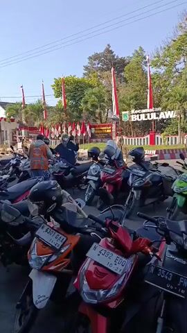 The video shows a busy outdoor scene with many parked motorcycles in the foreground. In the background, a building with a sign that reads "KECAMATAN BULULAWANG" is visible, adorned with red and white flags, suggesting a celebration or national event. Several people are standing around the motorcycles and near the building. One person in an orange vest appears to be directing traffic or managing the area. The weather appears to be sunny with clear skies. The motorcycles are a mix of colors and models, and they are parked closely together, indicating a significant gathering. The overall impression is one of a community event or a busy public space.