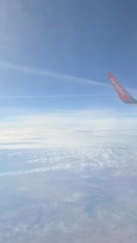 The video is an aerial view from an airplane window. The airplane wing, with the EasyJet logo visible, is in the right side of the frame. Below the plane, there is a layer of white clouds. Further down, faint patches of land are visible through gaps in the clouds. The sky is a clear blue with thin white contrails.