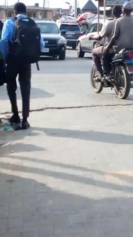 The video shows a street scene in a busy urban area. Two young men with backpacks, presumably students, walk along a sidewalk. A yellow van passes by. A black SUV is parked at the intersection. People walk across the street, some carrying items on their heads. There are motorcycles and other vehicles in the background. The setting appears to be a commercial area with buildings and overhead wires visible.
