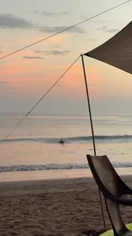 The video shows a beach scene at dawn or dusk. The sky is a mix of pink and pale blue. Calm waves are visible in the ocean, and a person is swimming in the water. In the foreground, part of a sunshade canopy and a black plastic chair are visible on the sandy beach. The camera zooms slightly toward the swimmer.