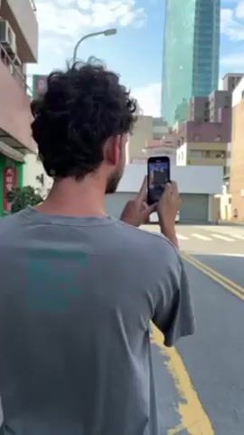 A man with dark curly hair, wearing a grey t-shirt, is standing on a city street taking a photo with his smartphone. The street has yellow lines marking the lanes. In the background, there's a tall green skyscraper and various other buildings of different heights and colors. The sky is blue with some scattered clouds. The man is facing away from the camera, and the phone screen is visible, showing the image he is capturing of the street and the buildings beyond. A white car is visible parked across the street.