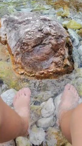 A first-person perspective shows two pairs of feet submerged in clear, shallow water. The water flows over smooth, multi-colored pebbles and larger, rougher rocks. One prominent, reddish-brown rock sits in the center foreground, partially submerged. The camera pans slightly to reveal a rocky riverbed with more pebbles and submerged rocks. In the background, a lush green forest lines a hillside, with a mountainous backdrop. Several people are visible scattered along the riverbank, some appearing to be sitting or wading in the water. The water is clear and appears to be a natural river or stream, possibly in a mountainous or forested region.