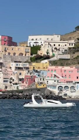 The video shows a colorful coastal town with a boat in the foreground. The town is built on a hill, and the buildings are painted in various bright colors such as yellow, pink, and blue. There's a church with a dome visible among the buildings. In the foreground, a white motorboat is moving on the water. The sky is clear and blue.
