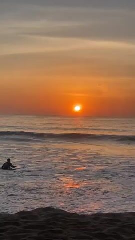 A serene beach scene unfolds at sunset. The sun, a bright orange orb, hangs low in the sky, casting a warm glow across the ocean. Gentle waves roll towards the shore, their crests breaking into white foam. The sand in the foreground is dark and textured. Two surfers are visible in the distance, their figures small against the vast expanse of the sea. The sky above transitions from a vibrant orange near the horizon to a soft grey, with wispy clouds creating subtle patterns. The overall atmosphere is tranquil and peaceful.