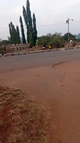 The video shows three people running on a street in an outdoor environment. The street is paved and has a cracked surface. The scene is set in a suburban-like area with trees and greenery visible in the background, along with utility poles and distant buildings. The camera is positioned at ground level, providing a slightly angled view of the street and the people running. Two of the people are wearing athletic clothing, while the third person is dressed in casual attire. The video appears to be captured during the day, with overcast lighting conditions.