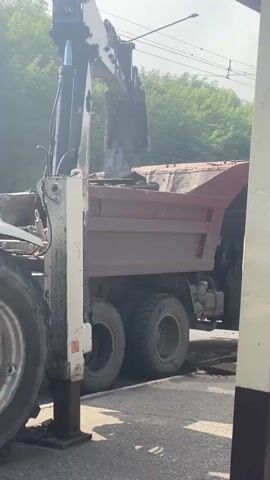 A backhoe is actively digging and loading material into the bed of a dump truck. The backhoe's arm is extended, and its bucket is tilted, depositing dirt into the truck. The dump truck is positioned alongside the excavation. The scene appears to be a construction or road repair site. Trees and utility poles are visible in the background. The lighting suggests it is daytime.