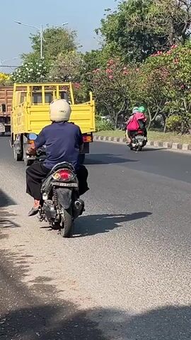 The video captures a street scene. In the foreground, a white car drives on the right side of the road, and a motorcycle is visible on the left side. In the middle ground, a yellow truck travels forward, followed by a motorcycle with two riders. The background features trees and buildings under a clear sky. The overall impression is a sunny day in an urban or suburban environment with ongoing traffic.