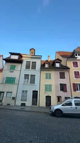 The video pans across a street lined with colorful buildings and parked cars under a clear blue sky. The buildings are of varying heights and architectural styles, with pitched roofs and multiple windows. The street is paved with cobblestones. A silver minivan is parked on the right side of the street, and a black car is parked further down on the same side. In the background, a white building with a tower and a steeple can be seen. The scene appears to be a quaint European town or village.