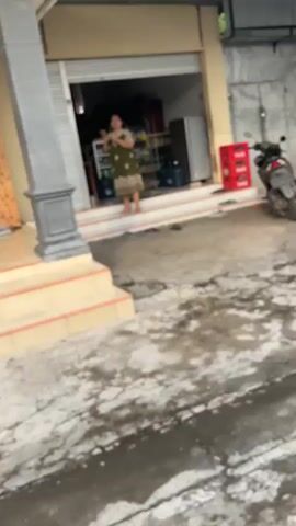 A woman in a green floral dress stands in the doorway of a shop. A motorcycle is parked to the right of the shop. The ground is paved with cracked concrete.